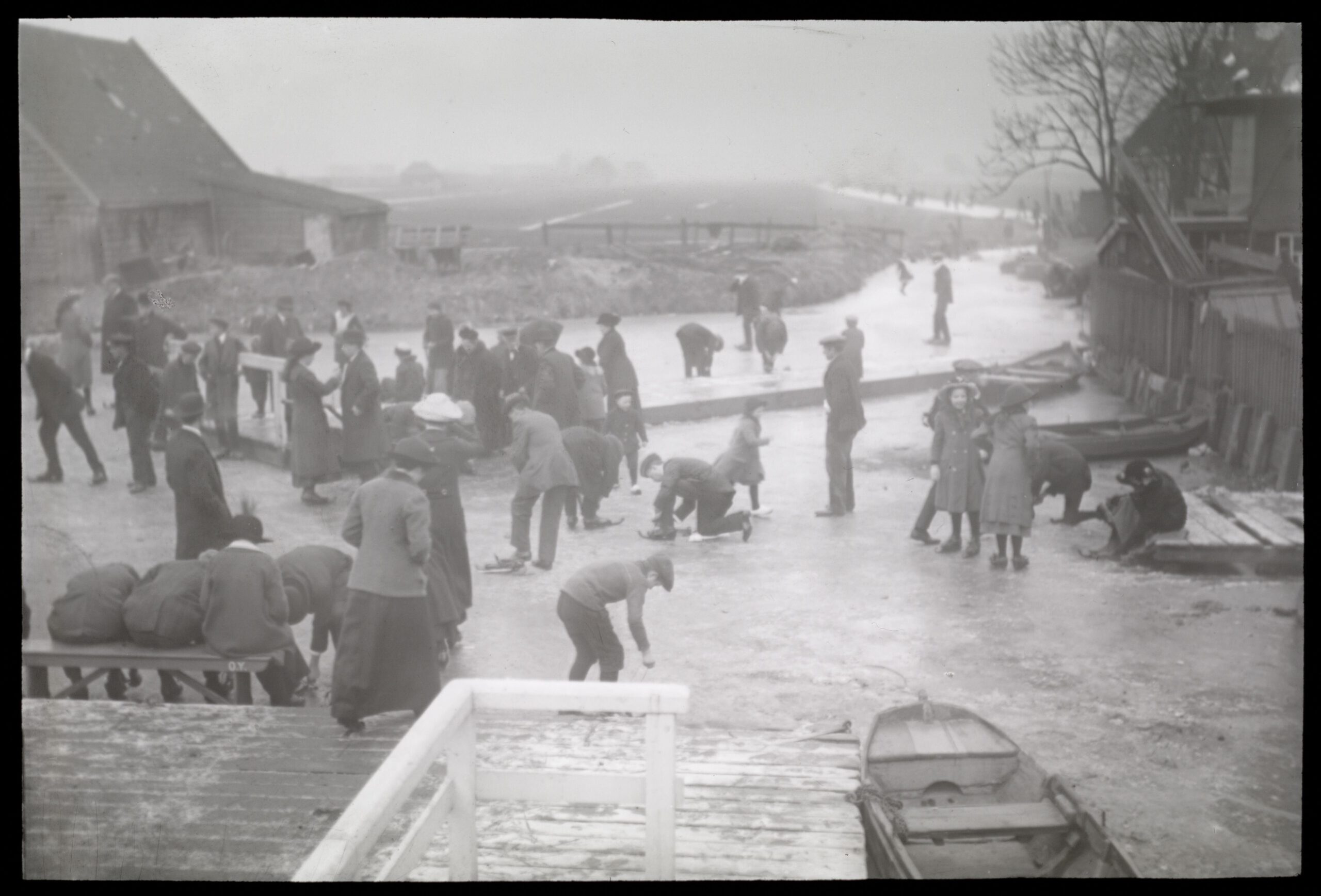 Lantaarnplaatje N116 uit de serie ‘Nederland in beeld en begrip’ – Een groep mensen op het ijs die hun schaatsen vastbinden. Op de achtergrond, tussen de weilanden, de Slotervaart waarop wordt geschaatst, thans Surinameplein. Gezien vanaf de steiger aan de Jacob Marisstraat.